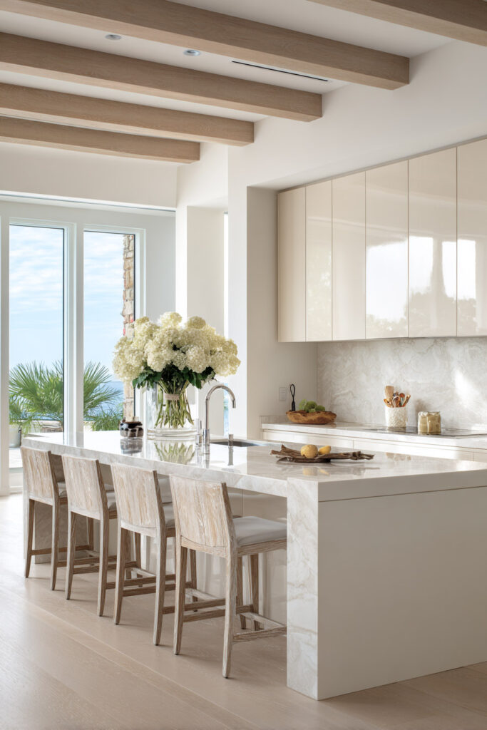 A bright neutral kitchen with a white marble island, light wood beams, glossy cream cabinets, and large glass doors opening to a coastal view.
