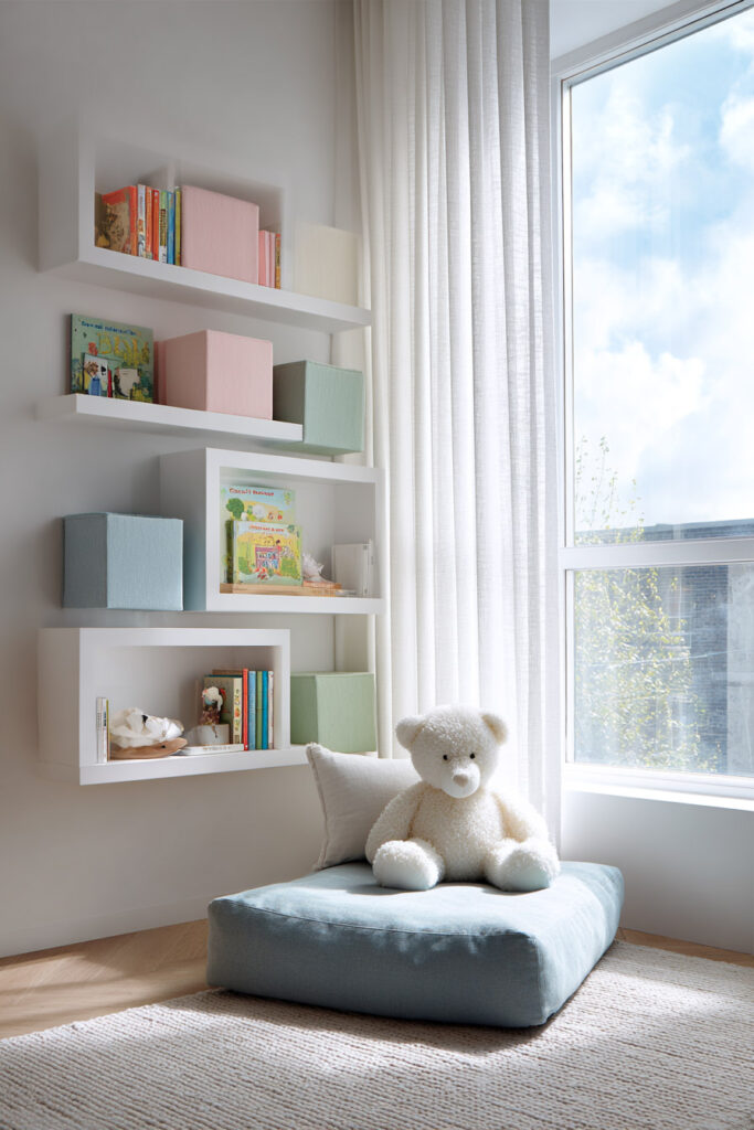 White wall-mounted kids’ room shelves with pastel storage bins, books, and a plush floor cushion with a teddy bear near a large sunny window.