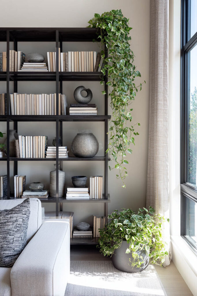 Tall dark wood bookshelf styled with neutral books, sculptural vases, and cascading greenery next to a light-filled window and soft gray sofa.