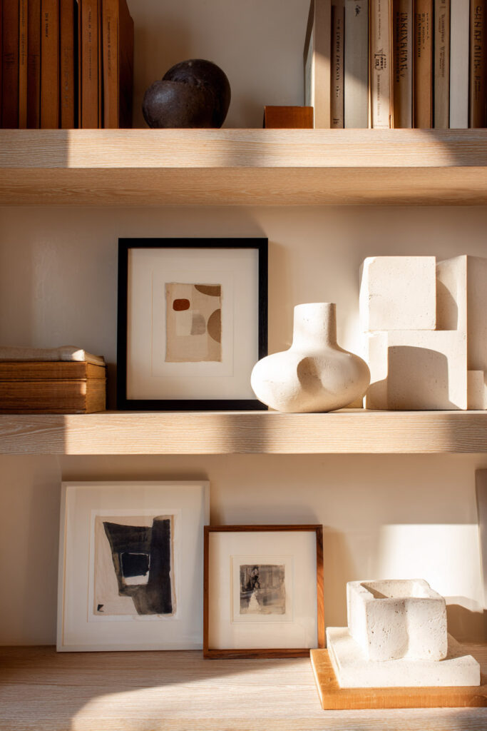Light wood shelves decorated with small abstract framed art, sculptural cream ceramics, stacked vintage books, and neutral-toned textures.