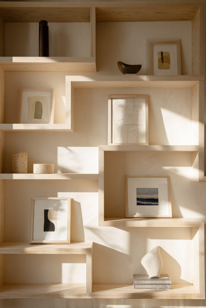 Built-in light wood shelves styled with minimalist framed art, sculptural objects, and soft neutral tones in a sunlit modern space.
