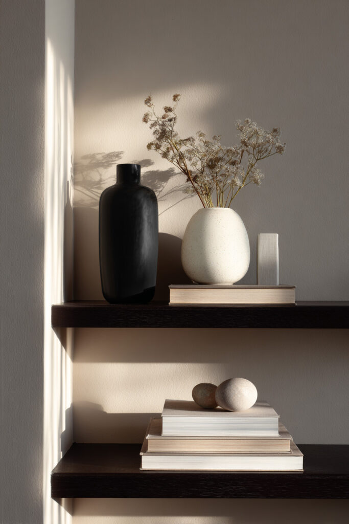 Dark wood floating shelves styled with stacked neutral books, a matte black vase, a speckled ceramic vase with dried flowers, and smooth sculptural stones, lit by soft natural light.