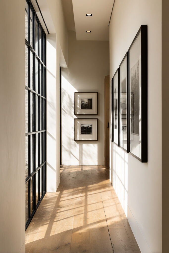 Minimalist neutral hallway featuring a gallery wall with black-framed monochrome artwork, light oak floors, and natural light streaming through tall black-framed windows.