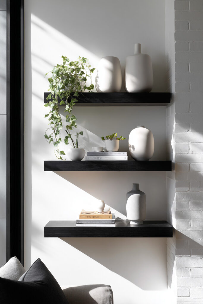 Three black floating shelves styled with neutral ceramic vases, stacked books, and a trailing ivy plant, set against a sunlit white wall with a modern aesthetic.