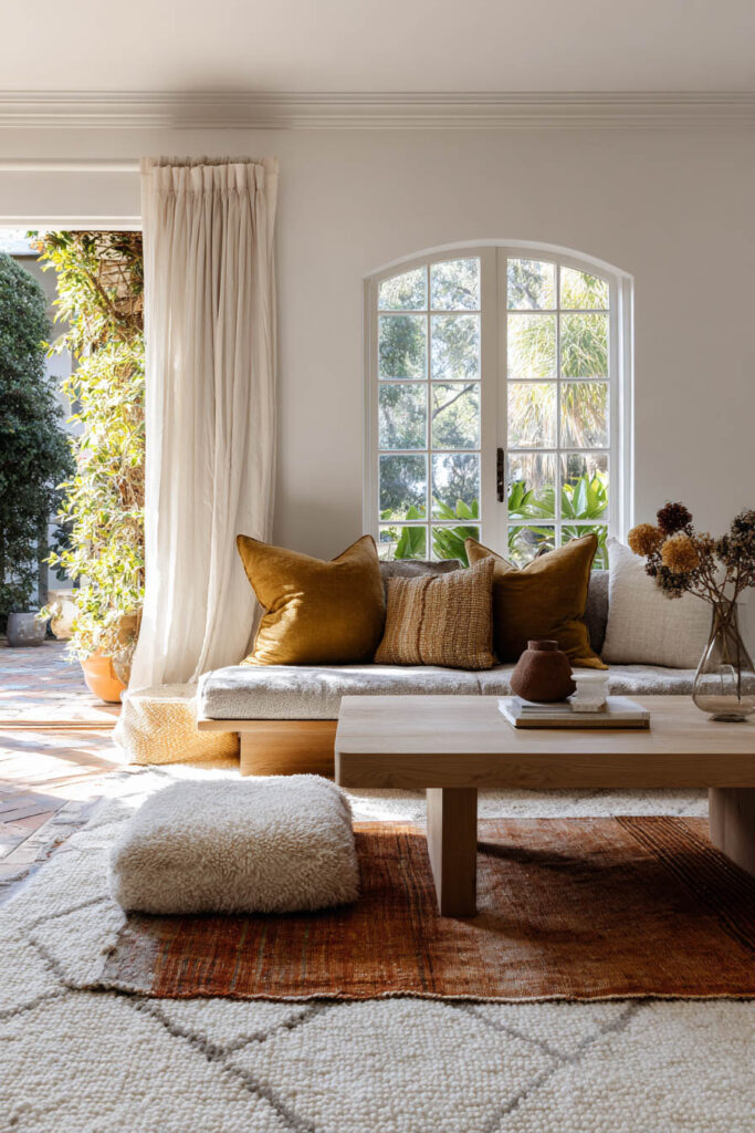 Bright fall living room with layered white and burnt orange rugs, a low wood coffee table, a built-in sofa topped with mustard and textured pillows, and soft sunlight streaming through French doors.