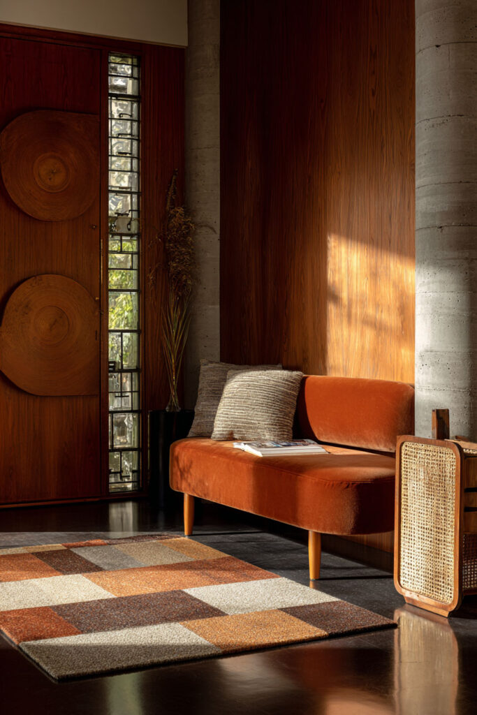 Mid-century style fall living room featuring a burnt orange velvet sofa, textured throw pillows, a checkered rug in warm autumn tones, and natural light streaming through tall windows.