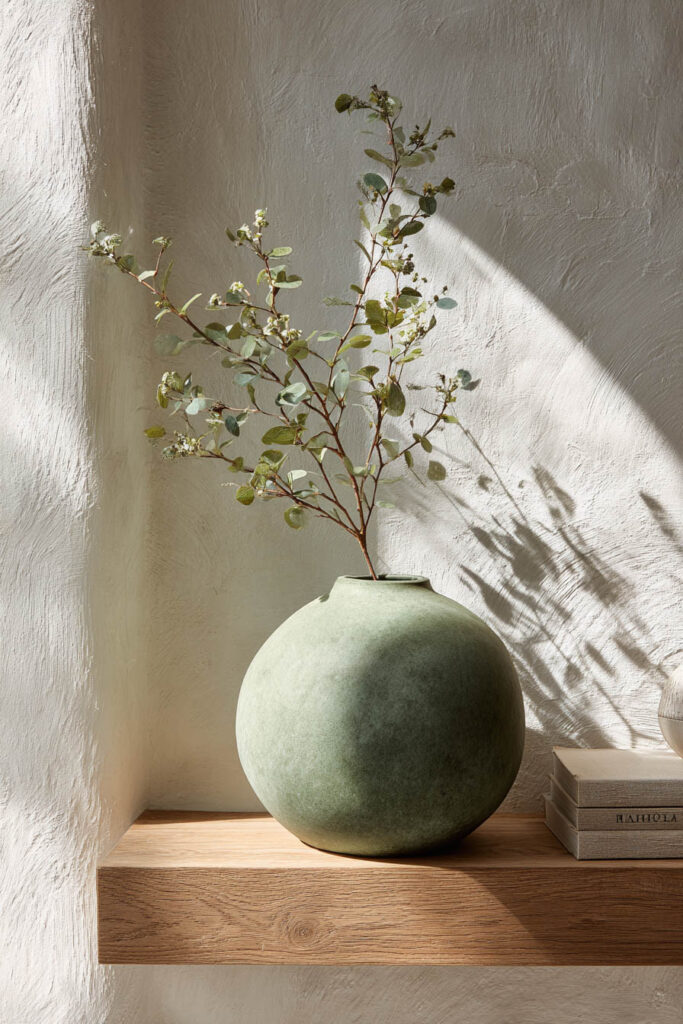Sage green round vase with eucalyptus branch styled on a wooden shelf against a textured wall, minimalist living room decor idea.