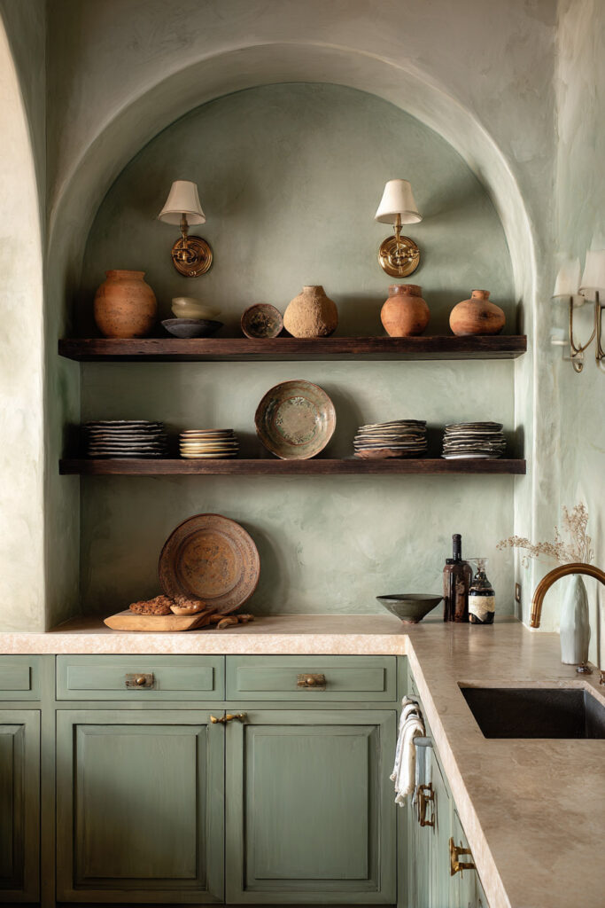 Elegant sage green kitchen with arched alcove, plaster walls, open wood shelves, brass sconces, and rustic pottery decor.