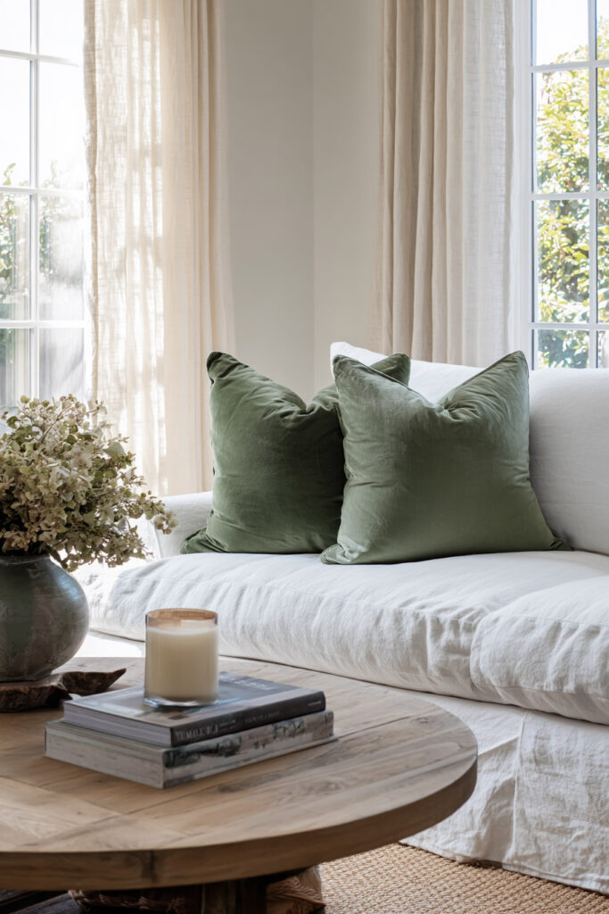 Living room with white linen sofa, sage green throw pillows, rustic wood coffee table, and vase of dried flowers in natural light.