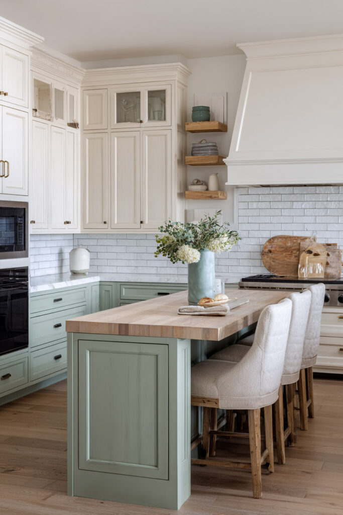 Farmhouse-style sage green kitchen with butcher block island, white cabinetry, brick backsplash, and upholstered barstools.