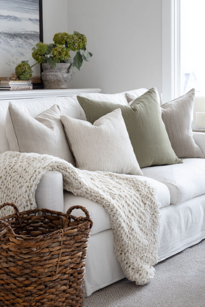 White sofa with sage green and neutral pillows, chunky knit throw, woven basket, and green hydrangea arrangement in living room.