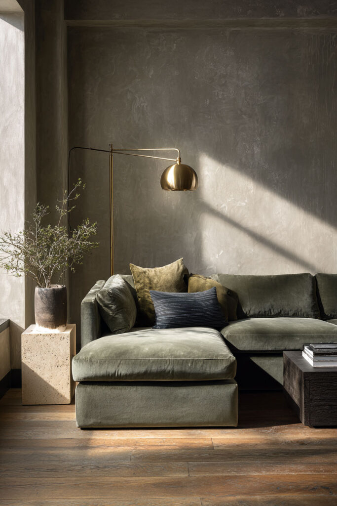 Moody sage green living room with velvet sectional sofa, brass floor lamp, stone pedestal planter, and textured plaster walls.