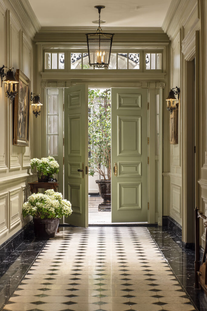 Grand sage green entryway with paneled double doors, checkered black-and-white floor, wall sconces, floral decor, and chandelier.