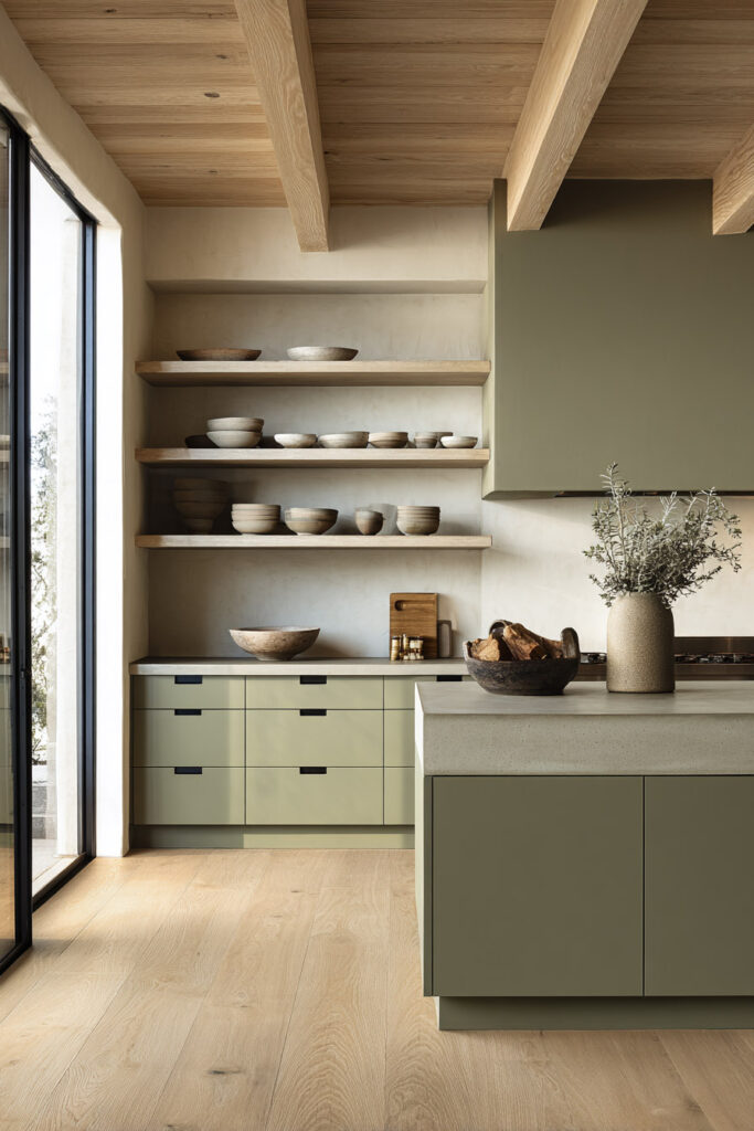 Modern sage green kitchen with flat-front cabinets, concrete countertops, open wood shelving, exposed beams, and large glass doors.