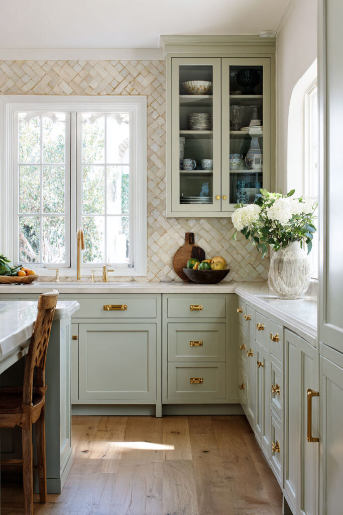 Sage green kitchen with brass hardware, glass-front cabinets, textured tile backsplash, and natural wood accents.