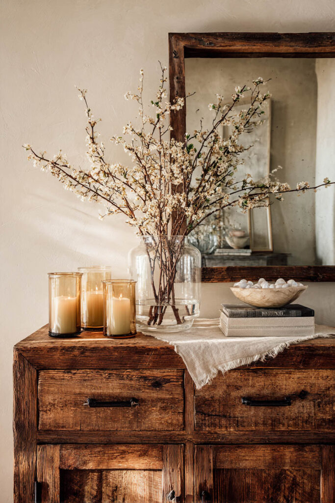 Rustic wooden console table with flowering branches in glass vase, amber candle holders, linen runner, and stone bowl decor.