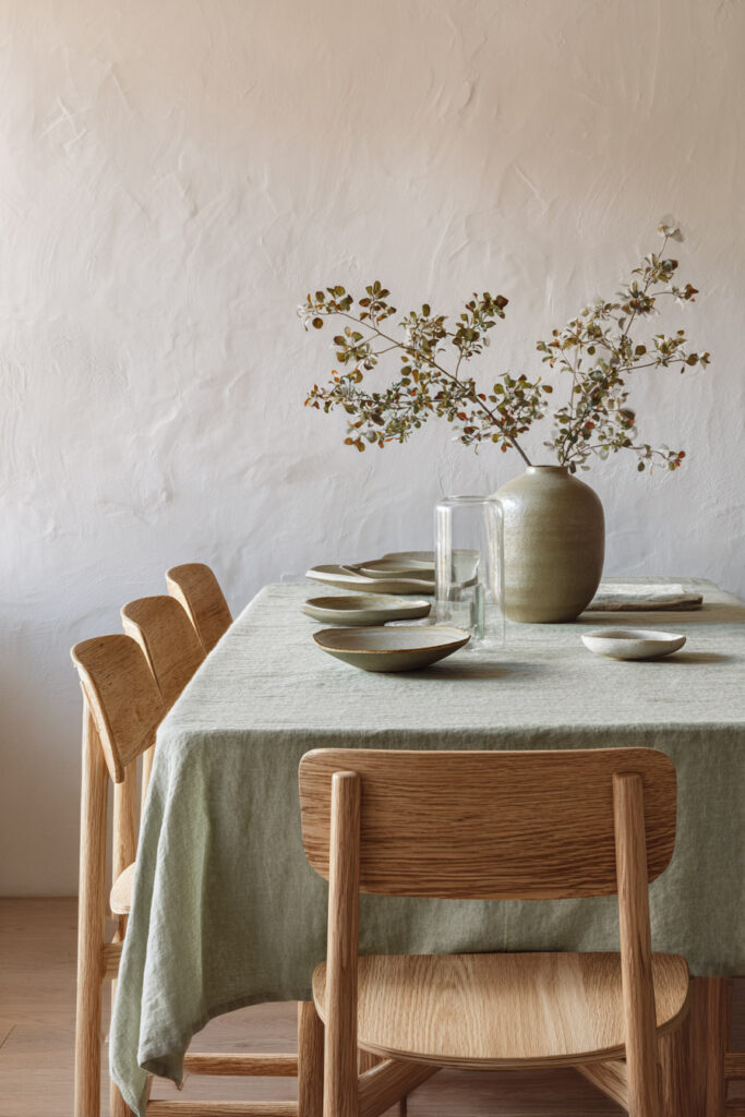 Minimalist dining room with sage green linen tablecloth, light wood chairs, ceramic tableware, and rustic vase centerpiece.