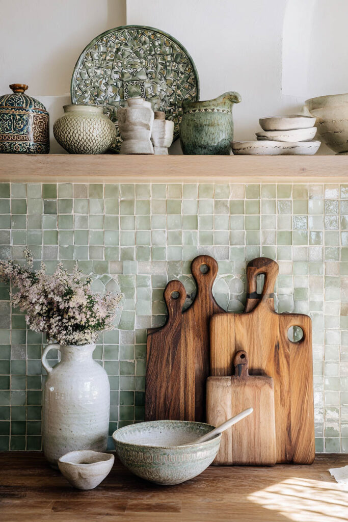 Sage green tiled kitchen backsplash with rustic wood cutting boards, artisanal pottery, and natural textures.