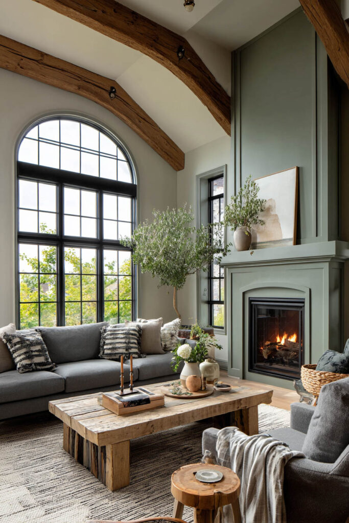 Rustic sage green living room with exposed wood beams, fireplace surround, arched windows, gray sofas, and farmhouse-style wood coffee table.