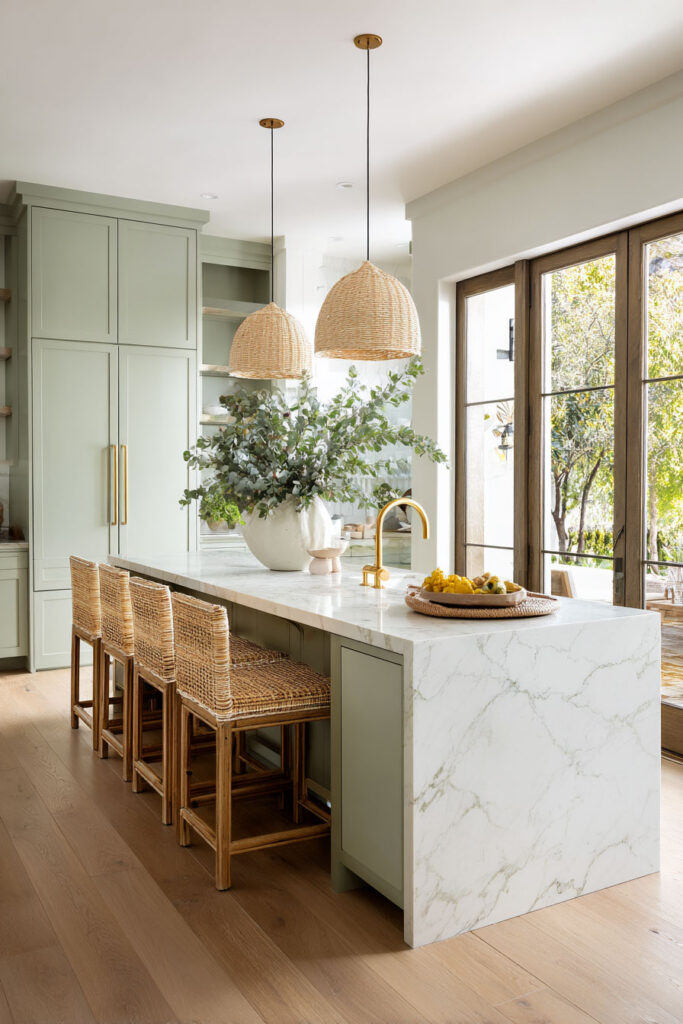 Sage green kitchen with marble waterfall island, woven rattan bar stools, basket pendant lights, and gold fixtures.