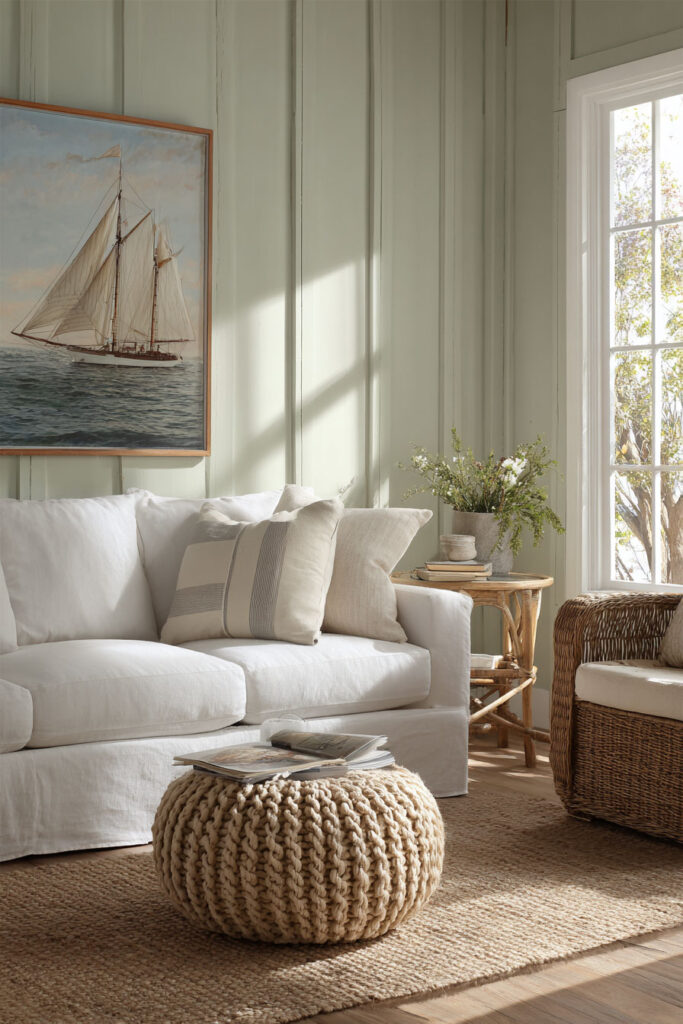 Coastal sage green living room with paneled walls, white slipcovered sofa, wicker chair, jute rug, and nautical artwork.