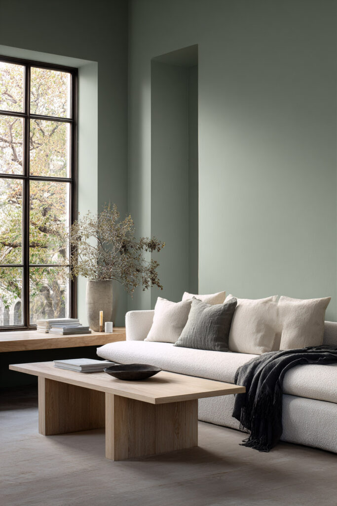 Minimalist sage green living room with cream sofa, neutral pillows, light wood coffee table, and large window with natural light.