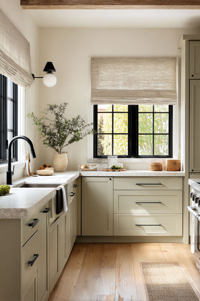 Light-filled sage green kitchen with black hardware, stone countertops, black faucet, Roman shades, and natural wood flooring.