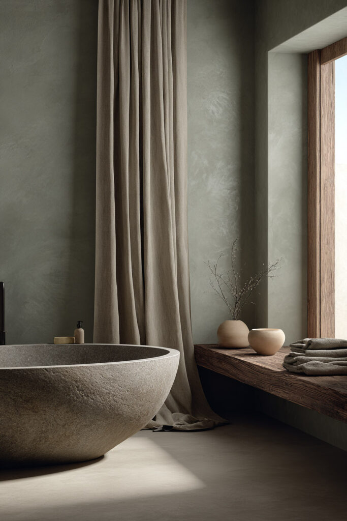 Sage green bathroom with stone soaking tub, natural wood bench, linen curtains, and ceramic accents for a serene spa-inspired retreat.