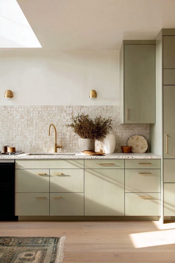 Sage green kitchen with brass hardware, terrazzo countertops, stone backsplash, and modern cabinetry in a bright, natural light setting.