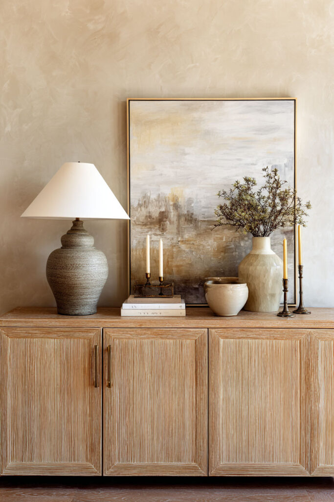 Natural wood sideboard styled with sage green ceramic vase, abstract artwork, candlesticks, and earthy living room decor accents.