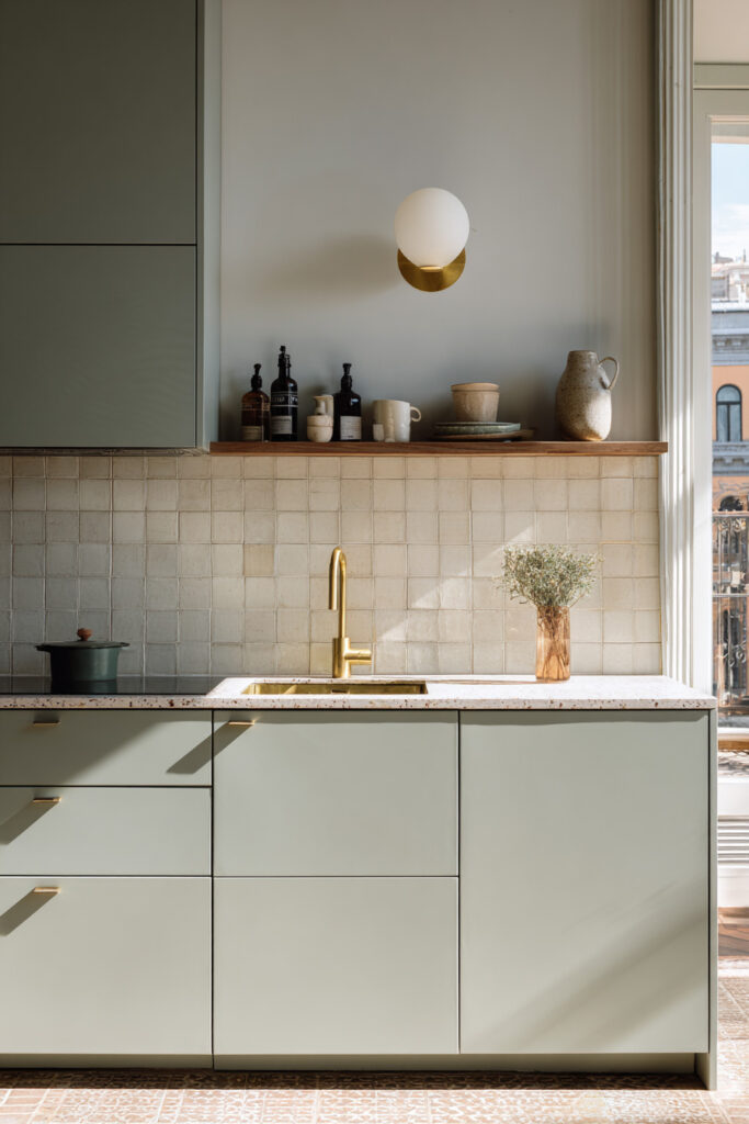 Sage green kitchen with brass faucet, modern cabinetry, tile backsplash, wooden shelf, and natural light.