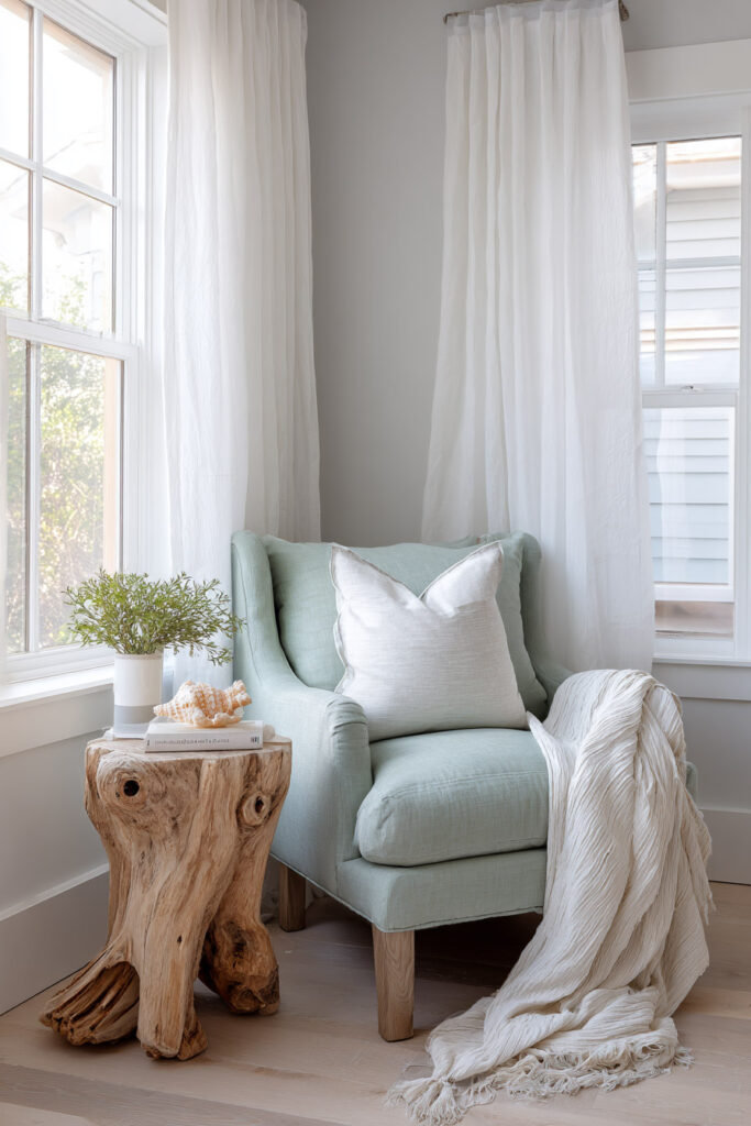 A cozy coastal reading nook with a sage green armchair, driftwood side table, white throw blanket, and soft natural light filtering through sheer white curtains.