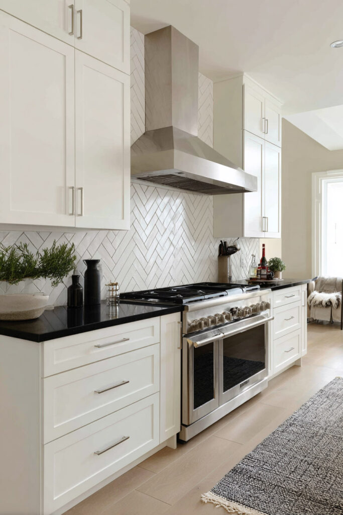Black and white kitchen with white shaker cabinets, black countertops, stainless steel range, herringbone backsplash tile, and natural wood flooring.