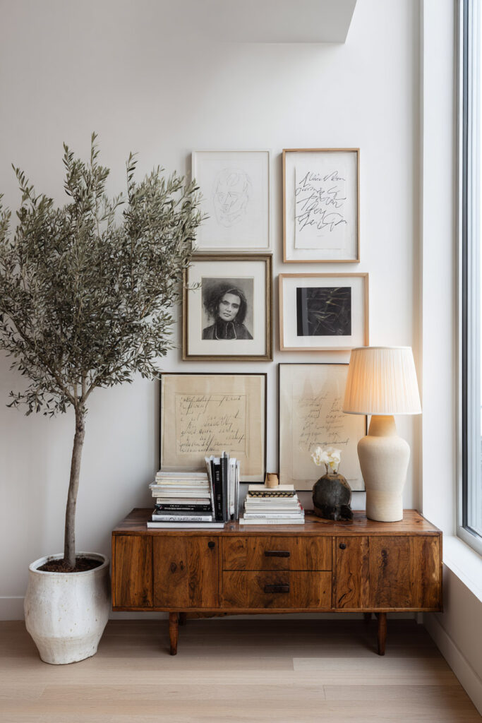 Minimalist gallery wall with sketches and handwritten art above mid-century wood console table, olive tree, and ceramic lamp.