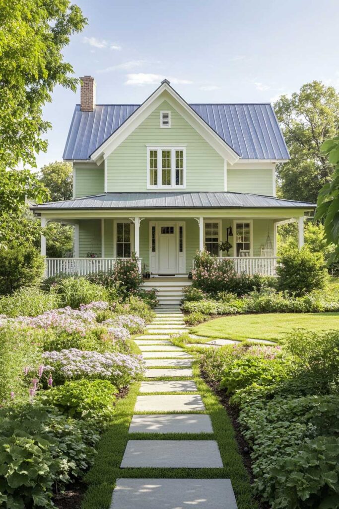 Light sage green farmhouse with blue metal roof, white trim, wraparound porch, and blooming garden pathway surrounded by greenery.