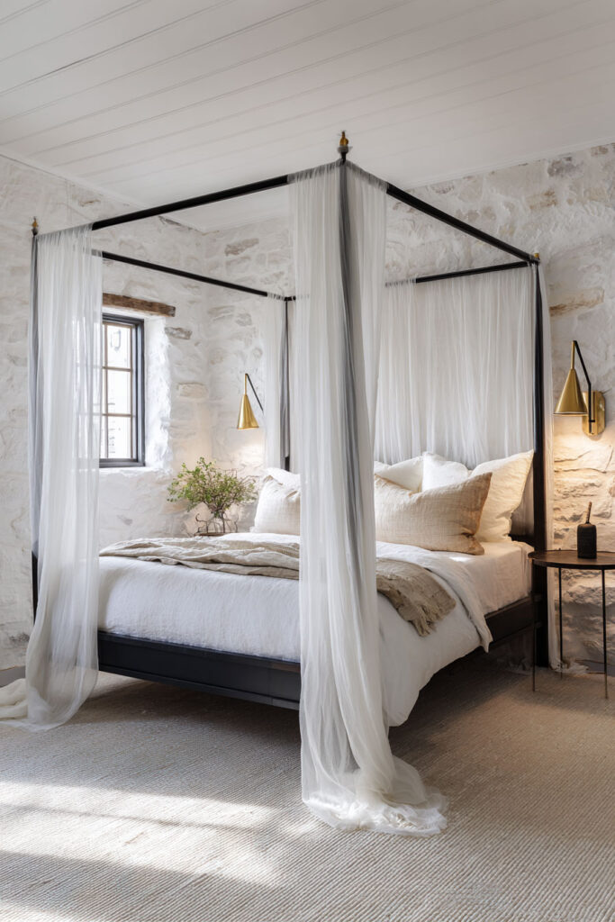 Minimalist black and white bedroom with stone walls, sheer canopy bed, and layered neutral bedding in a cozy rustic-modern style.