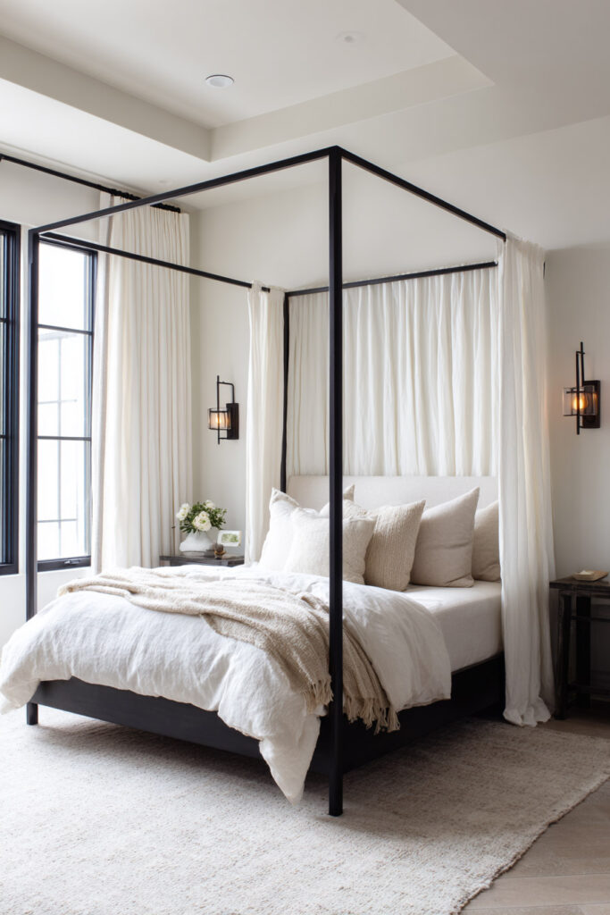 Cozy black and white canopy bed with sheer drapes, layered neutral bedding, and soft filtered light through modern black-trimmed windows.