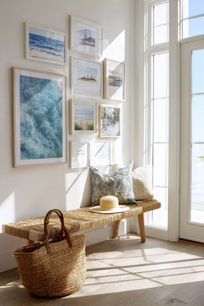 Coastal entryway with beach photo gallery wall, woven bench, straw tote, and blue accent pillows in bright natural light.