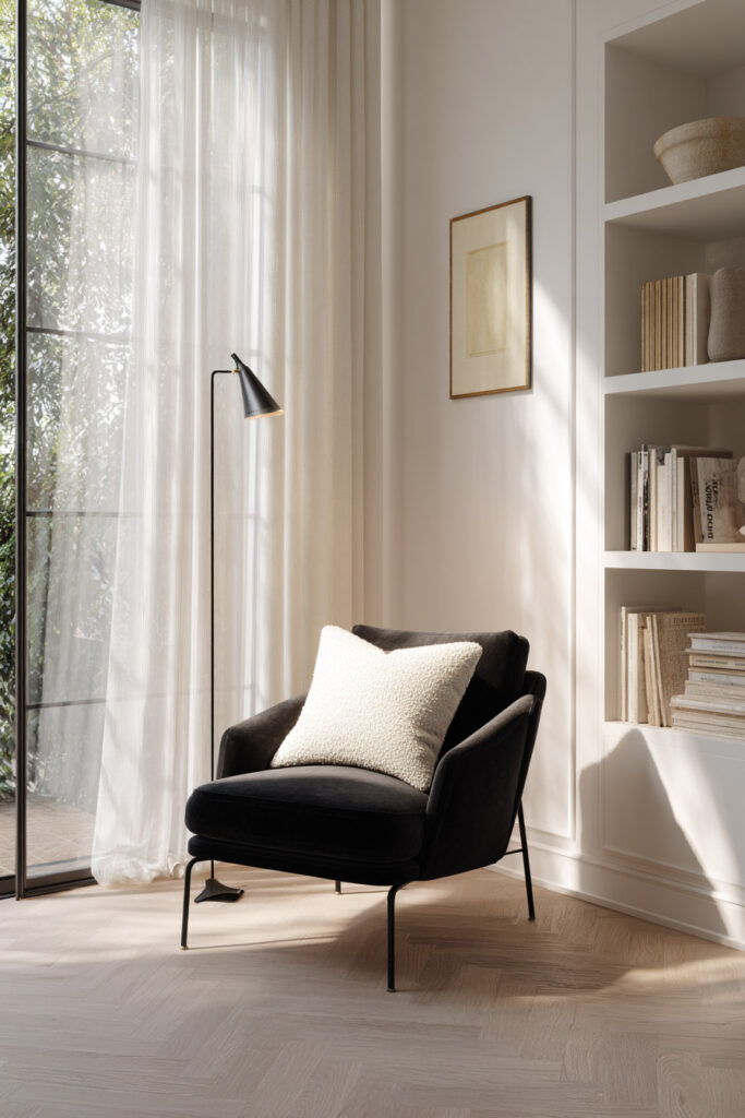 Cozy black and white reading nook with a black armchair, white boucle pillow, floor lamp, built-in bookshelf, and soft natural light through sheer curtains.