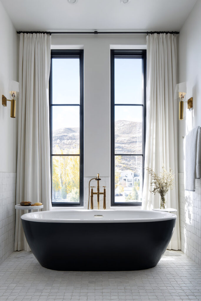 Elegant black and white bathroom with freestanding black bathtub, brass fixtures, white tile floors, and tall black-trimmed windows framed by flowing curtains.