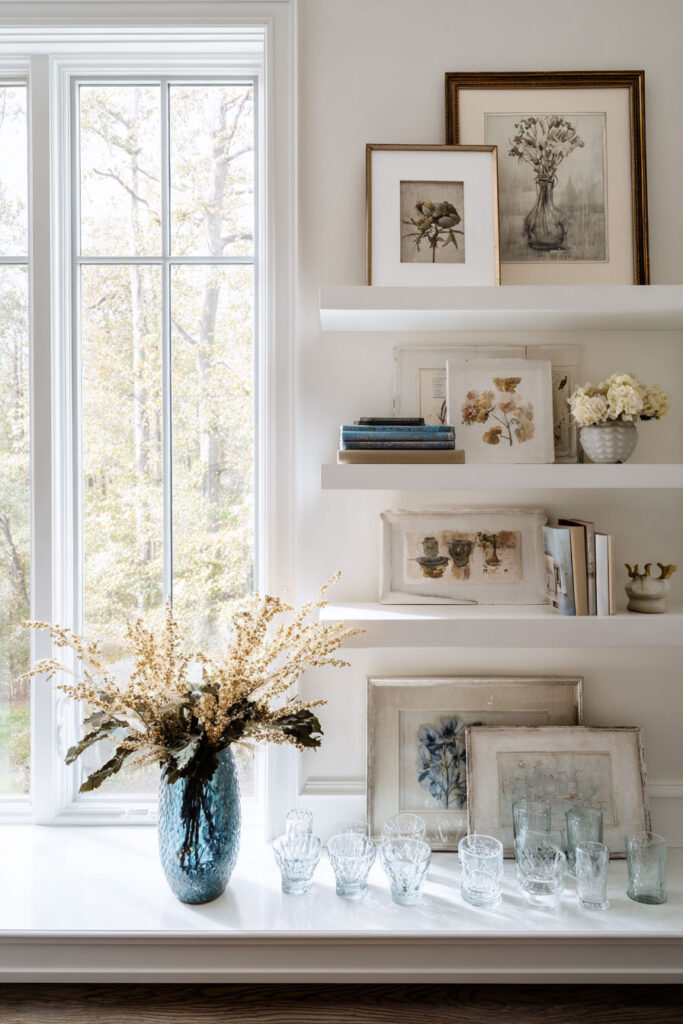 Floating shelves styled with vintage floral art prints, glass vases, books, and neutral seasonal decor beside bright window.