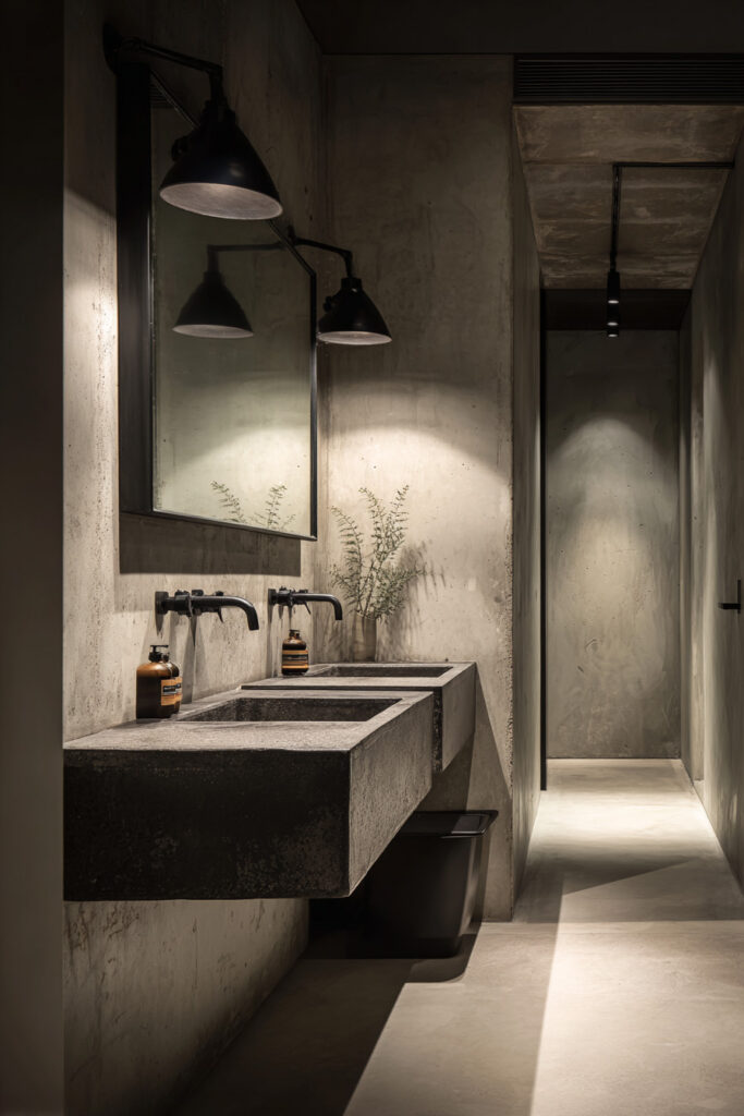 Minimalist monochrome bathroom with concrete walls, double trough sink, matte black fixtures, and moody lighting design.