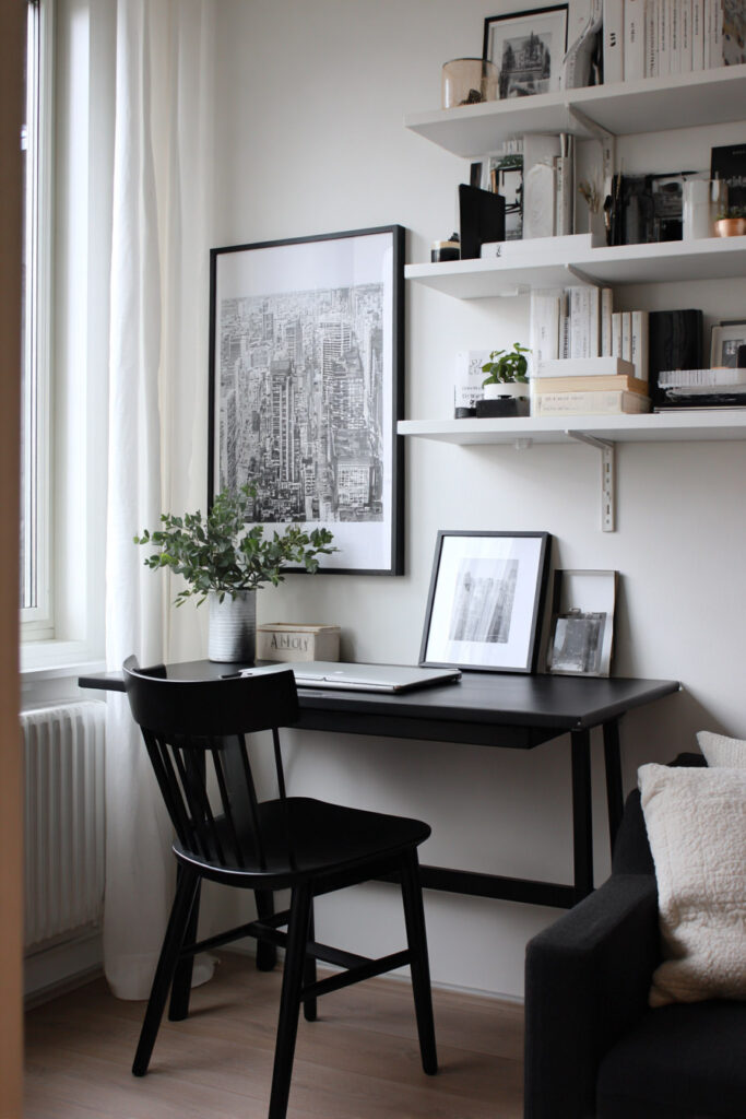 A minimalist black and white home office with a compact black desk, black chair, white wall shelves, and framed black-and-white artwork. Light wood floors and sheer curtains soften the space, creating a bright and cozy modern work nook.