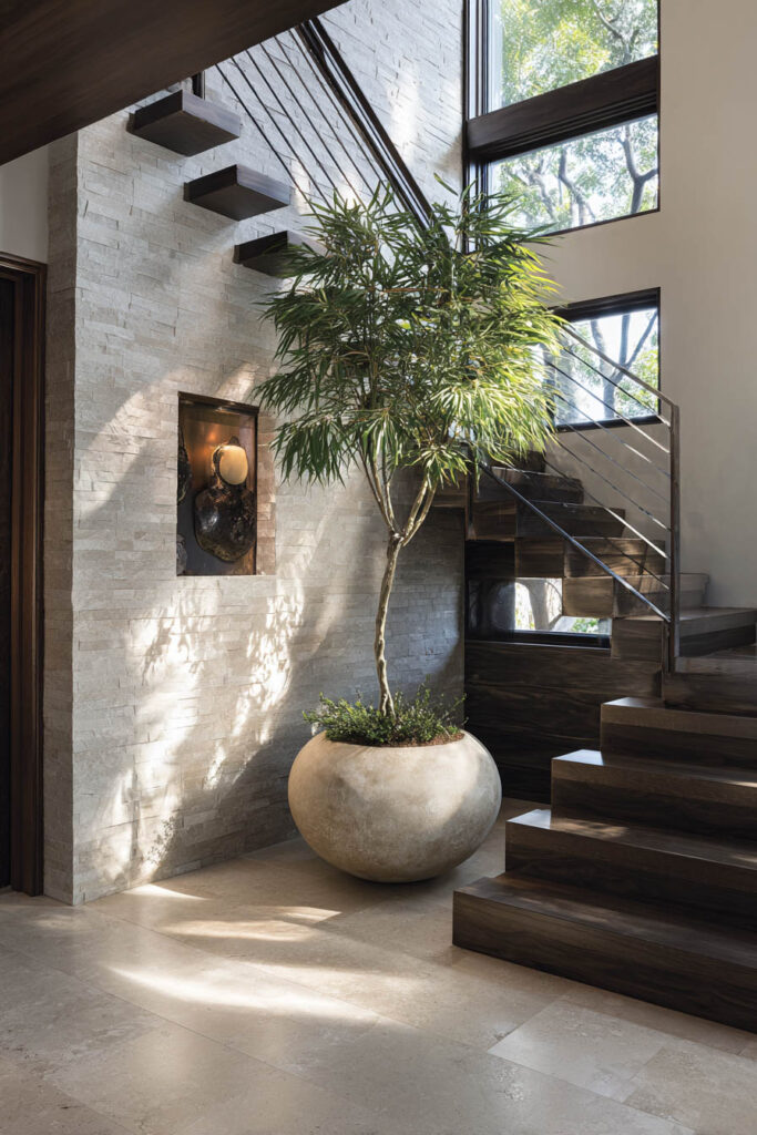 Modern staircase with tall indoor tree in stone planter beside textured wall and large windows with natural light.