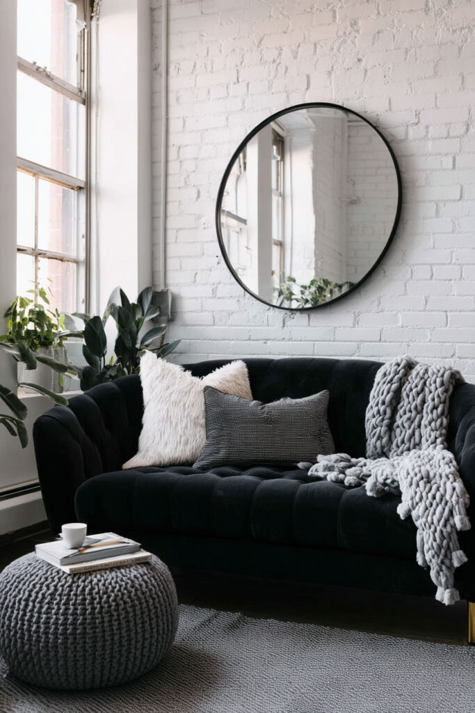 Industrial-style black and white living room with a black velvet sofa, round wall mirror, white brick wall, and cozy textured pillows and throws.