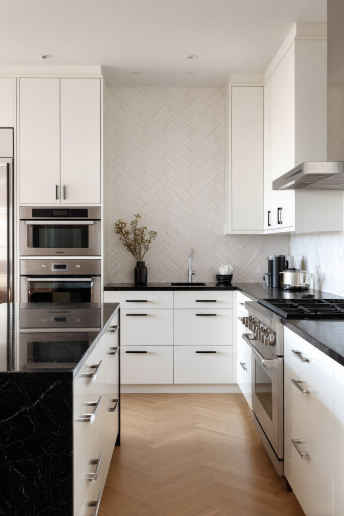 Modern black and white kitchen with white cabinetry, black countertops, herringbone tile backsplash, stainless steel appliances, and a black marble island.