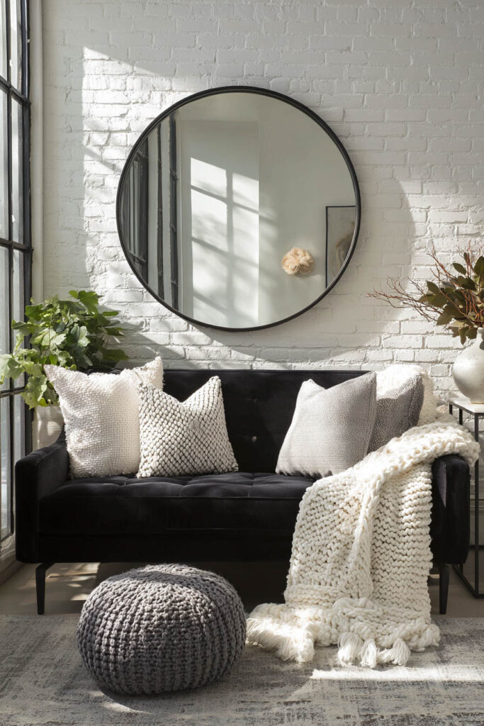 Black and white living room corner with black velvet sofa, white brick wall, textured throw pillows, round mirror, knit throw, and gray pouf in natural light.