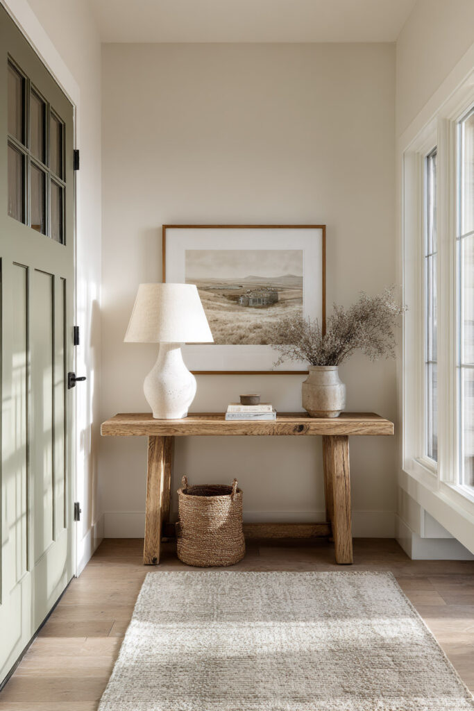 Neutral entryway with sage green door, rustic wood console table, ceramic lamp, woven basket, and dried floral arrangement in natural light.
