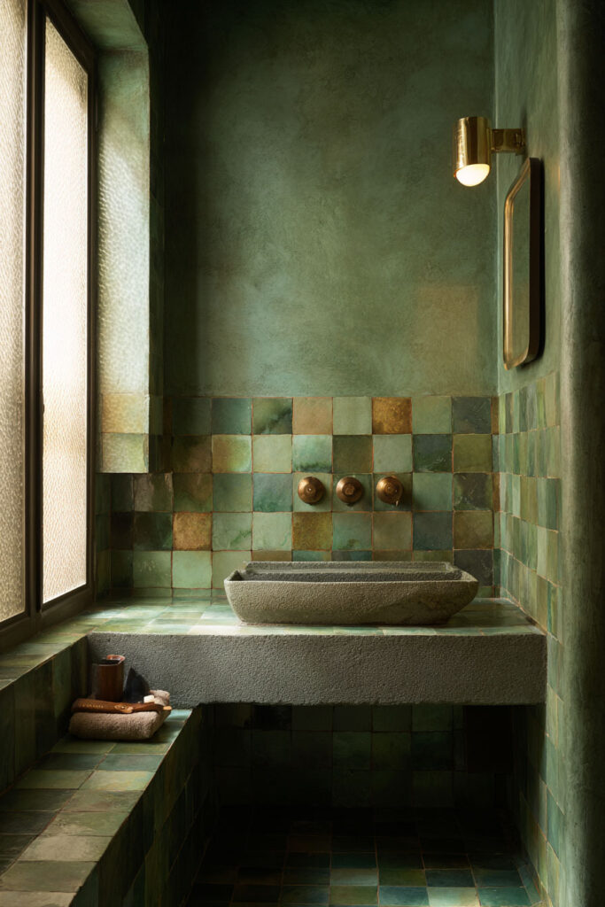 Earthy green bathroom with handmade tile walls, stone basin sink, brass fixtures, and natural light creating a warm organic color palette.
