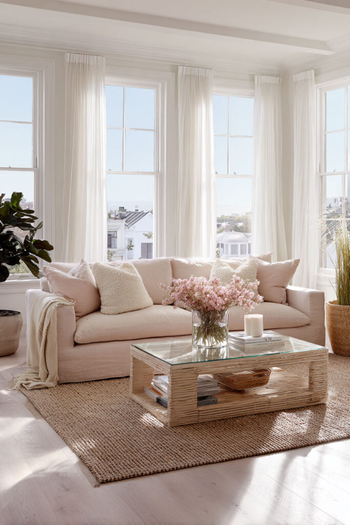 Light and airy living room with blush sofa, glass coffee table, woven rug, and natural sunlight streaming through white curtains.